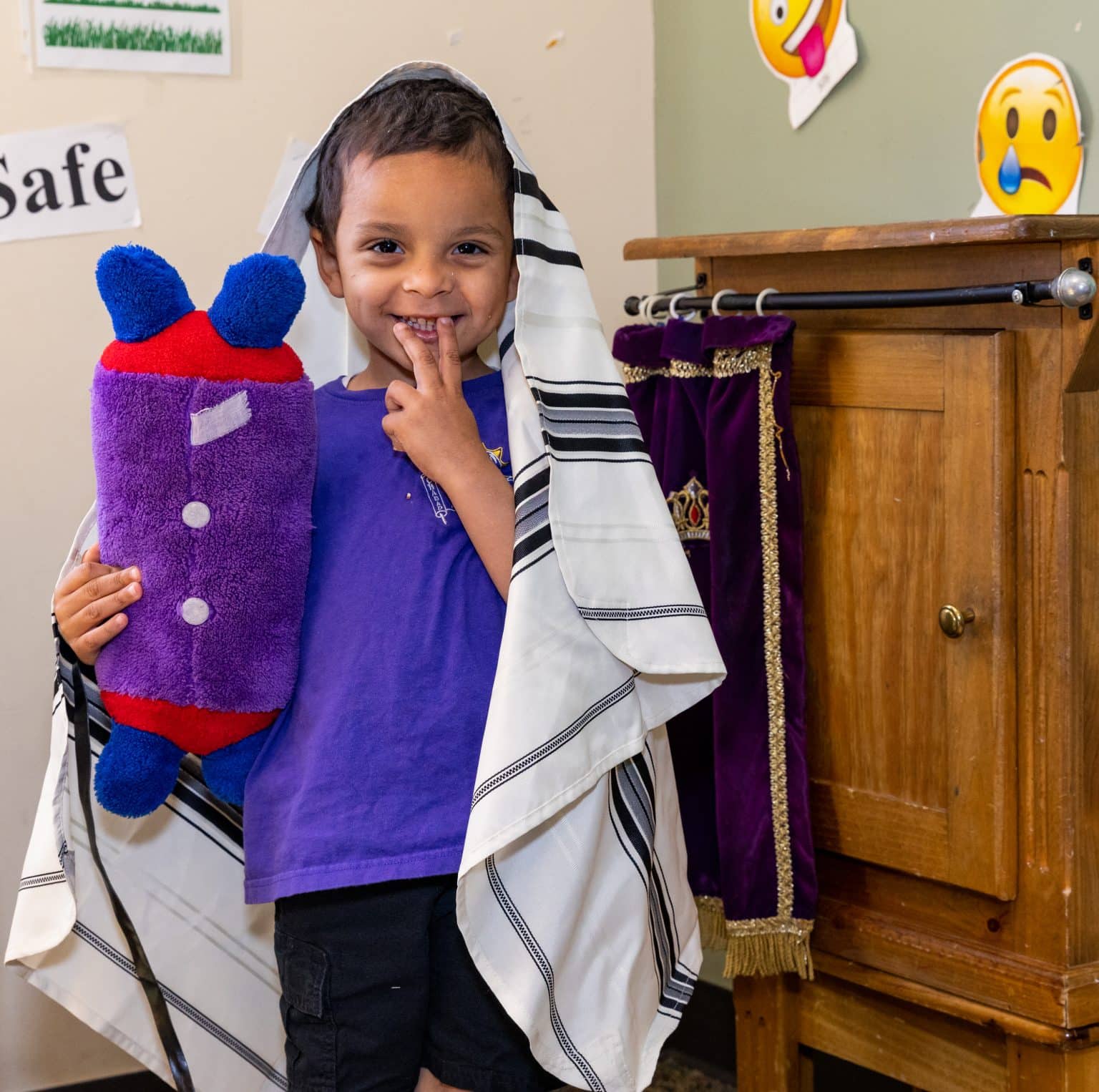 A smiling boy wearing a tallit holds a purple Torah plushie, with "Safe" on the wall behind him.