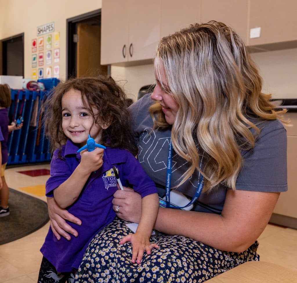 A smiling young girl in a purple shirt holds a blue toy while sitting on a blonde woman's lap.