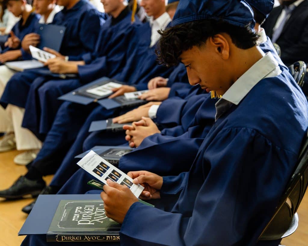 A young man in a blue graduation gown and cap looks down at a booklet, with "Pirkei Avot" on a book.