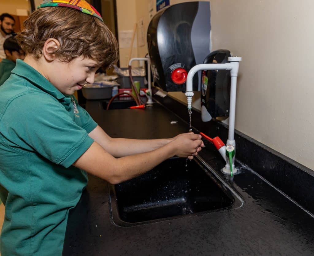 A boy in a green polo and kippah washes his hands under a faucet in a black sink.