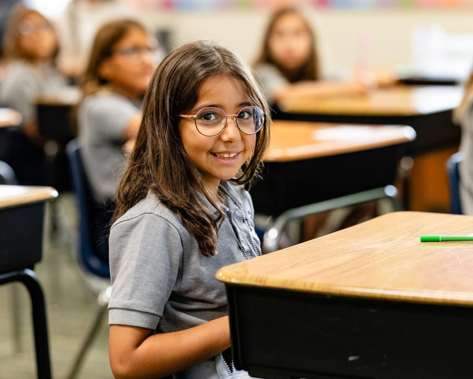 A smiling girl with long brown hair and glasses sits at a school desk in a classroom.