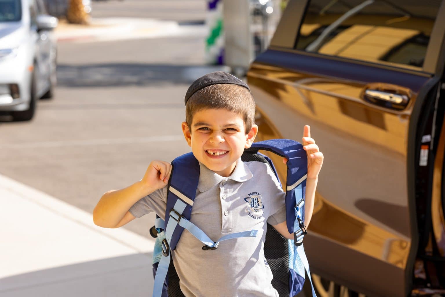 A smiling boy in a yarmulke and grey polo shirt with a blue backpack stands by an open car door.
